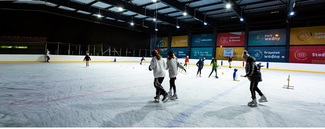 Zdjęcie przedstawia grupę osób jeżdżących na łyżwach po Oleśnickim Lodowisku ATOL Ice Rink. Lodowisko jest zadaszone i oświetlone.